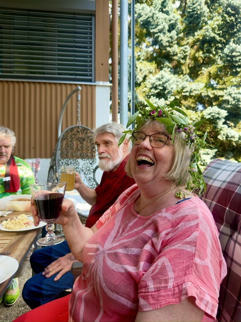 Elfi lacht mit Blumenkranz beim Fest, Hermann im Hintergrund